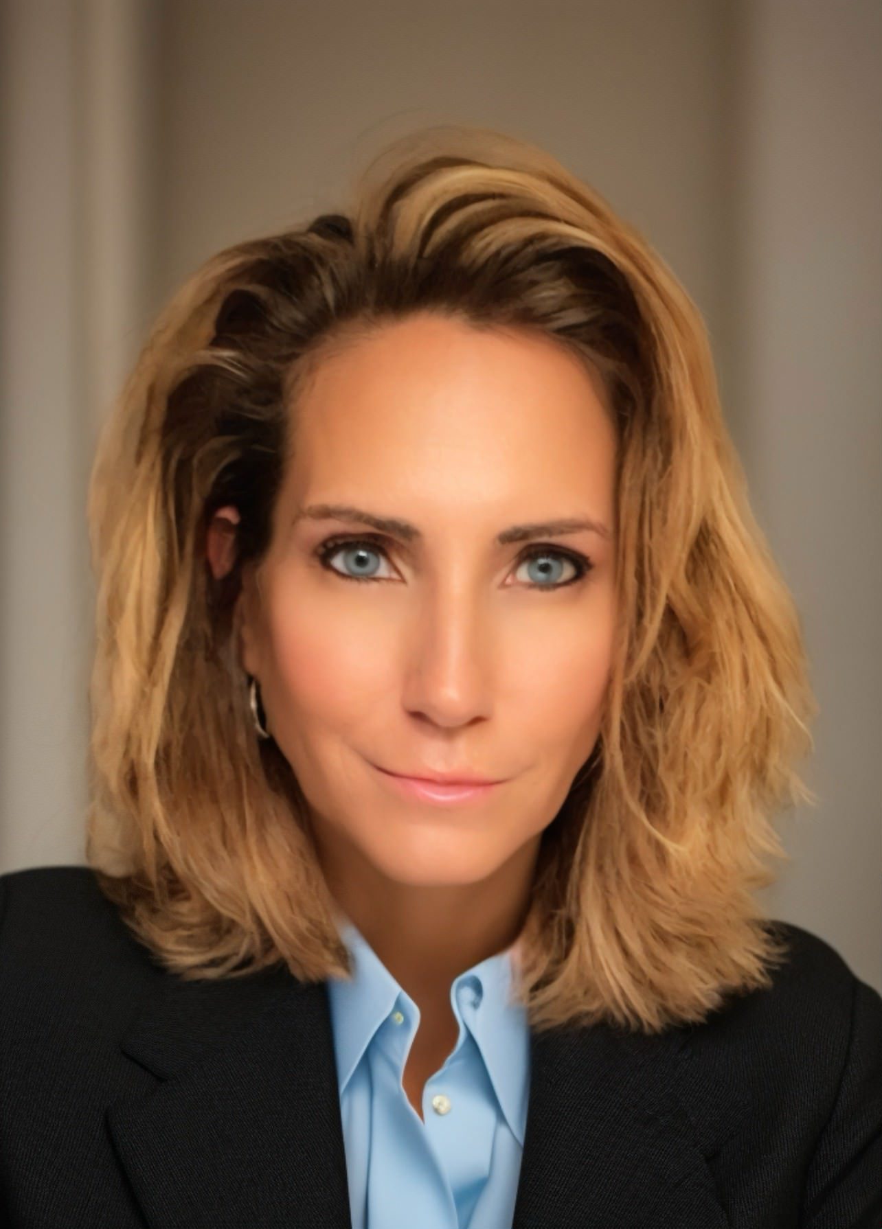 Portrait of a person with shoulder-length light brown hair, wearing a blue shirt and black blazer, looking directly at the camera with a neutral expression.