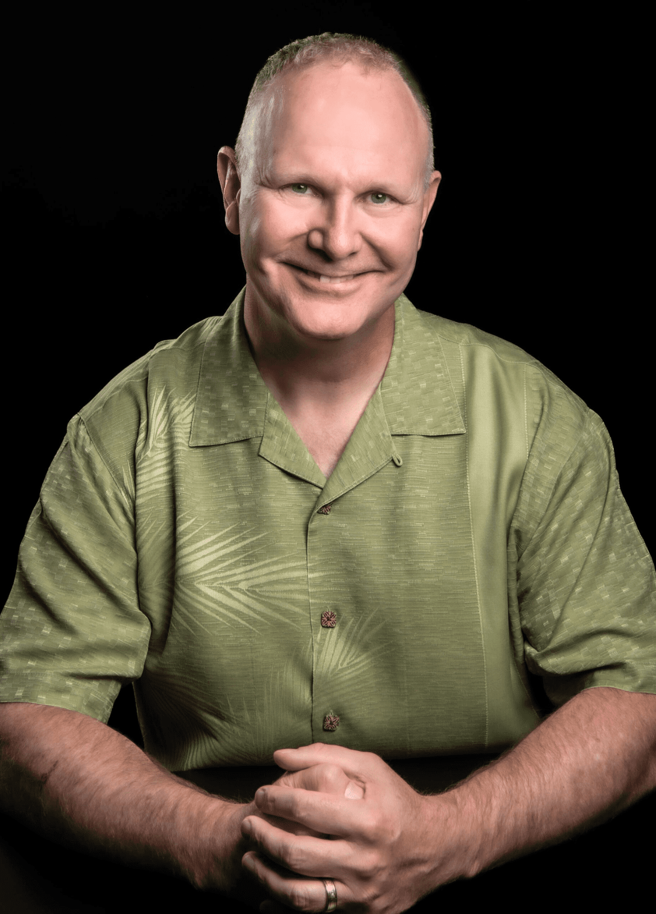 A smiling bald man sits with his hands clasped on a dark surface, wearing a light green collared shirt, against a black background.